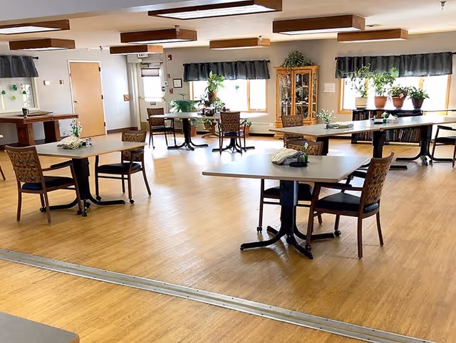 Bright communal dining room with several tables and chairs, potted plants, and wood-look flooring.
