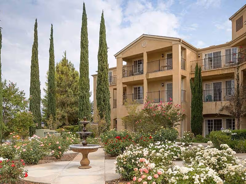 Exterior view of a multi-story senior living facility building with balconies, surrounded by a landscaped garden featuring tall cypress trees, flowering bushes, and a central stone fountain.