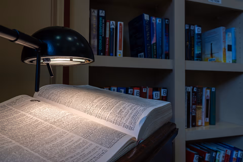 A close-up view of an open book illuminated by a black desk lamp, with bookshelves filled with various books in the background.