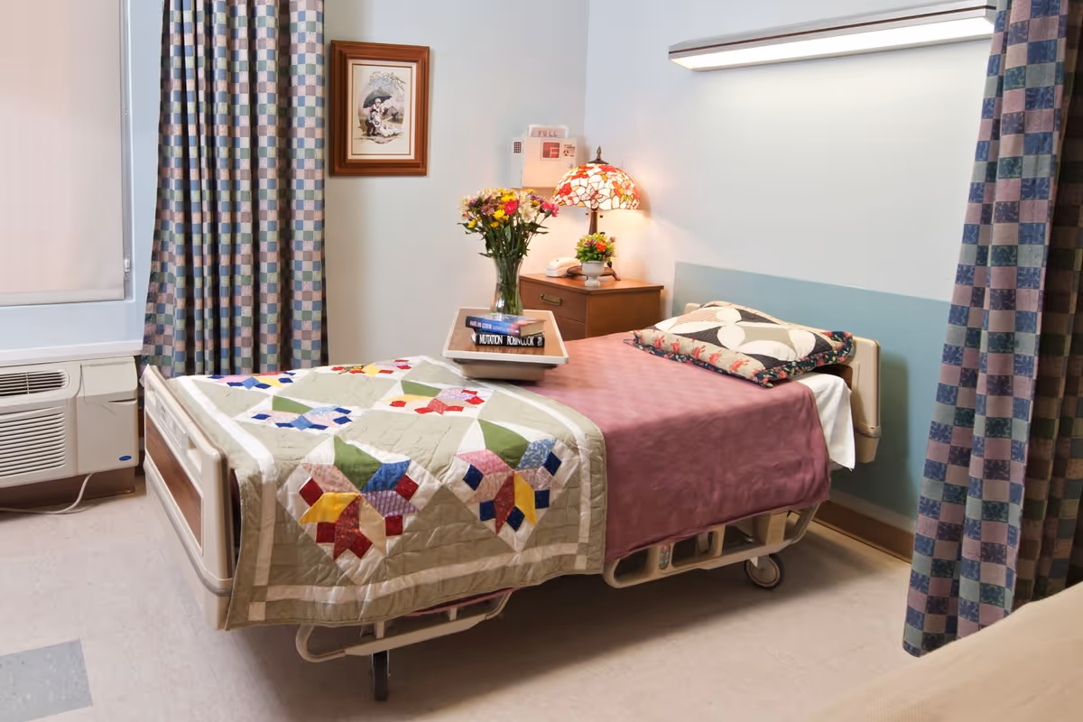 A neatly made hospital-style resident bed with a colorful quilt, bedside table with a lamp and flowers, and patterned curtains in a senior living bedroom.