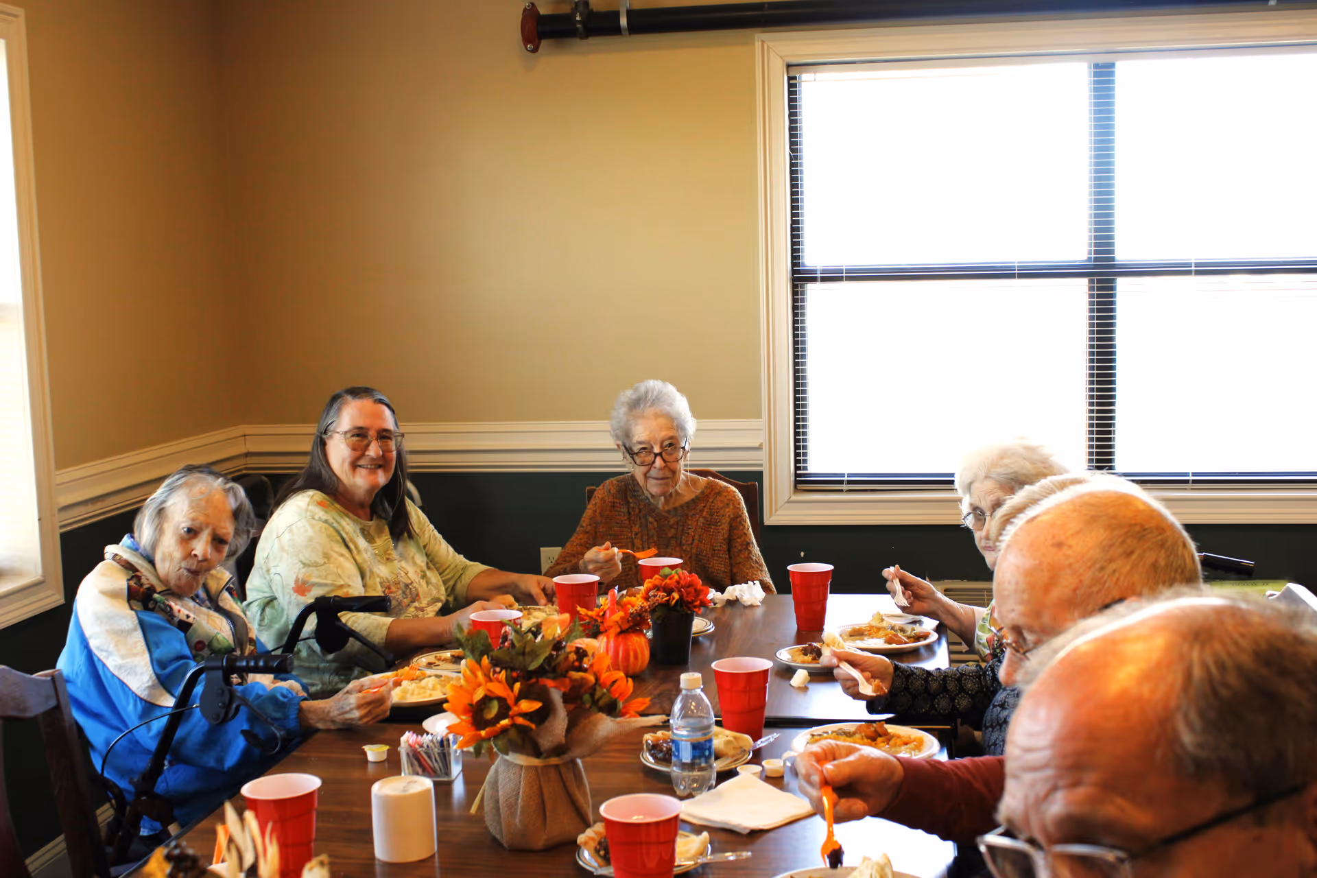 A group of elderly residents sitting around a dining table eating a meal with a fall-themed centerpiece.