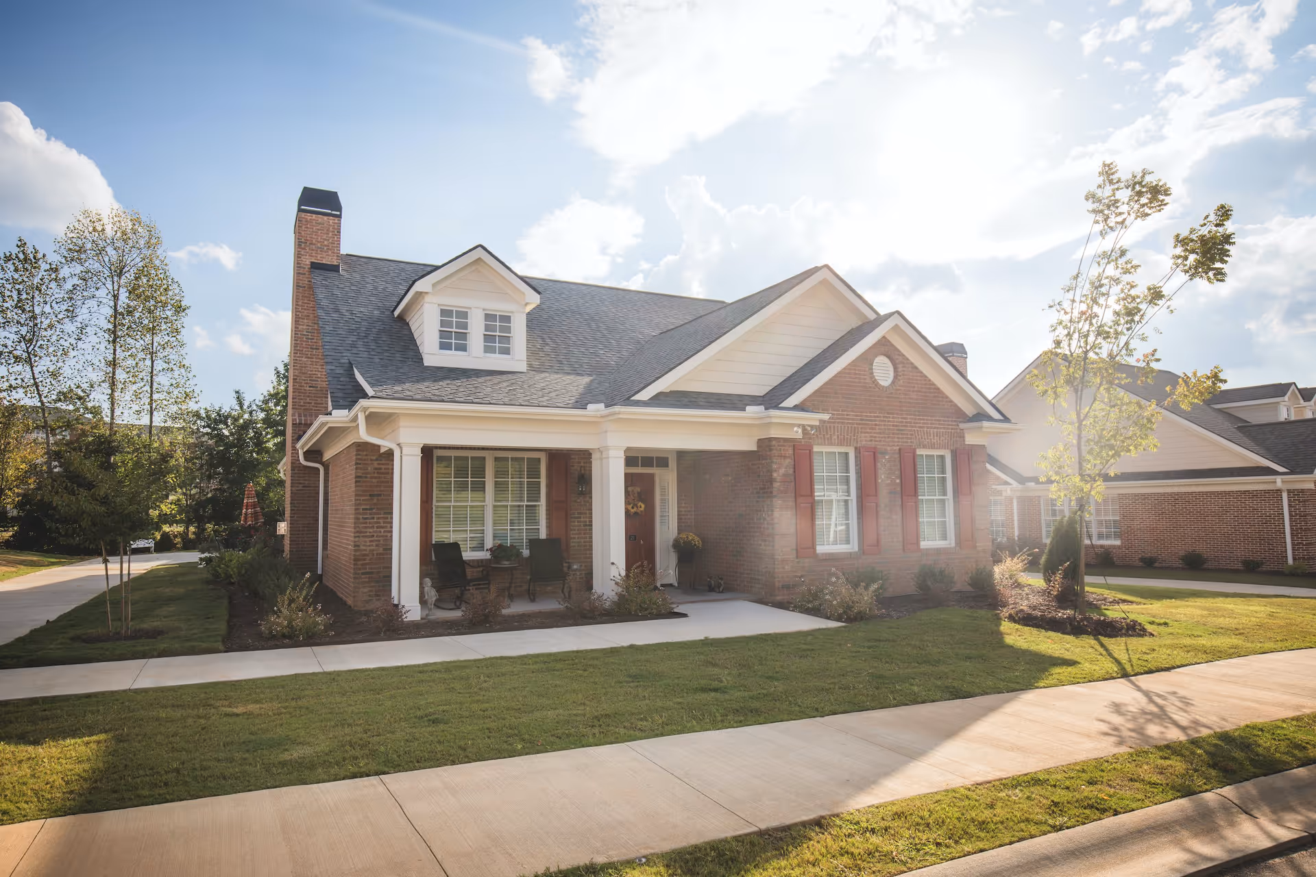 A single-story brick house with a gray shingled roof, white trim, and red shutters. The house has a small covered porch with two chairs and a table. There is a well-maintained lawn and a concrete sidewalk in front of the house. The sky is partly cloudy with sunlight shining through.