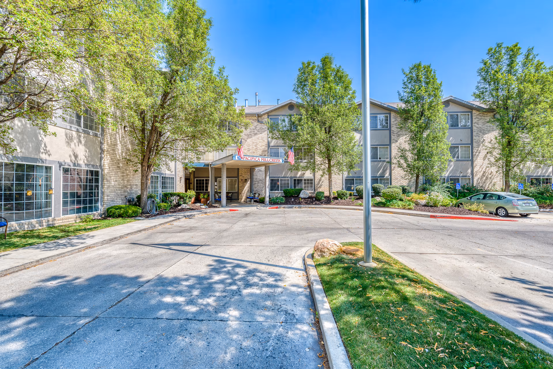 Exterior view of a senior living facility building with a driveway and entrance canopy. The building is three stories tall with large windows and surrounded by trees and landscaping. A car is parked near the entrance, and the sky is clear and blue.