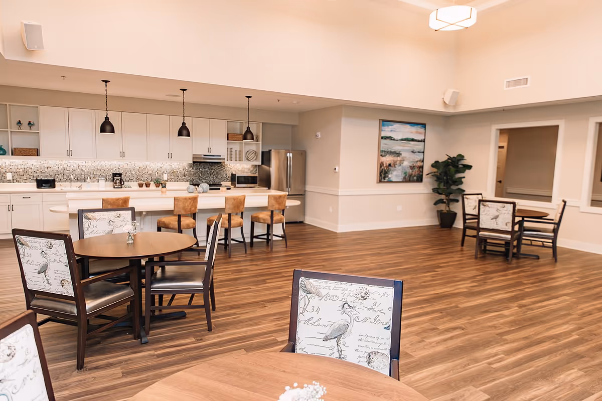 A spacious dining area with wooden floors, round tables with chairs featuring bird and script designs, and a modern kitchen in the background with white cabinets, a mosaic tile backsplash, stainless steel refrigerator, and three pendant lights hanging over a breakfast bar with four brown cushioned stools. A large painting and a potted plant are visible on the far wall.