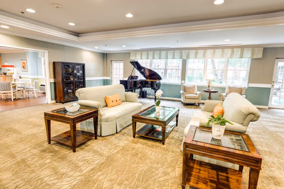 A bright and spacious living room area in a senior living facility featuring two beige sofas with orange accent pillows, three wooden glass-top tables with decorative plants, a grand piano near large windows, and two armchairs with a small round table and lamp between them. The room has beige carpet flooring, soft green and beige walls, and recessed ceiling lights. In the background, a dining area with tables and chairs is visible through an open doorway.