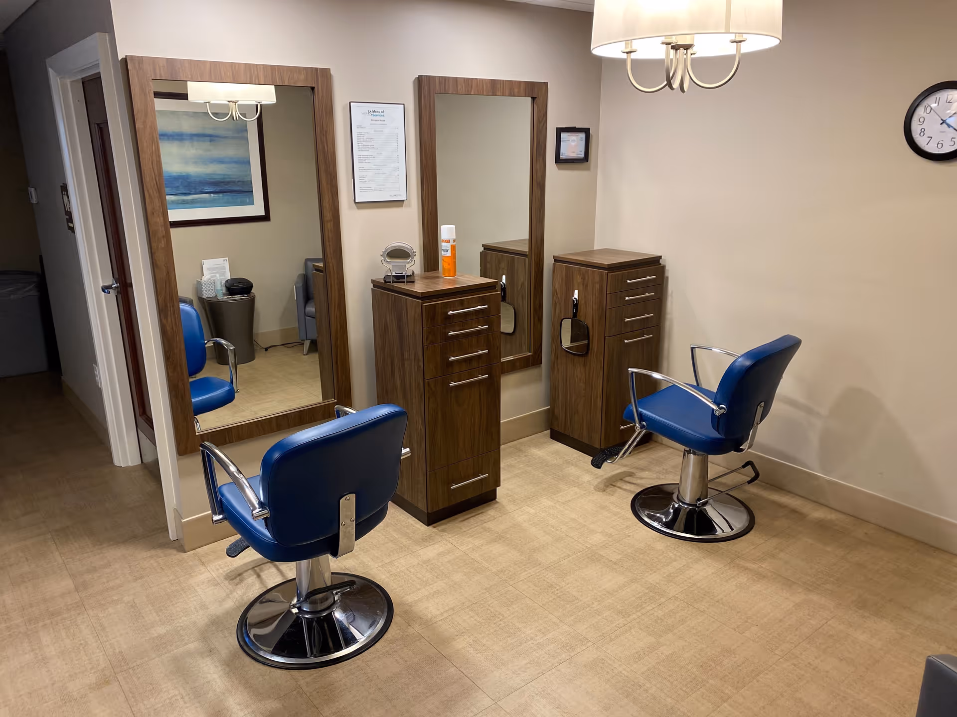 Interior view of a hair salon area in a senior living facility with two blue salon chairs facing large wooden-framed mirrors mounted on the wall. Each station has a wooden cabinet with drawers and a small mirror attached. The room has beige walls, a clock on the wall, and a ceiling light fixture.