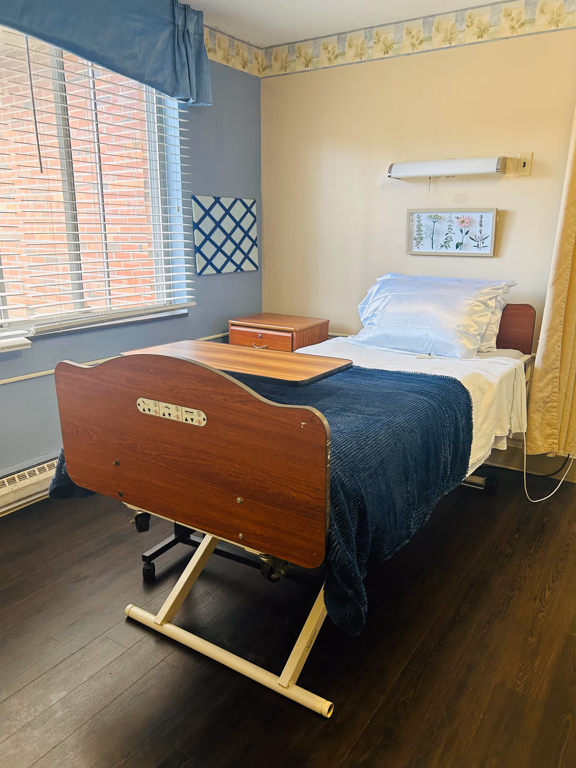 A single hospital-style bed with white sheets and a dark blue blanket in a room with wooden flooring. The bed has an adjustable overbed table and a wooden headboard and footboard. There is a small wooden nightstand next to the bed, a window with blinds and a blue valance, and a framed botanical print on the wall above the bed. The walls are painted in light yellow and blue with a floral wallpaper border near the ceiling.