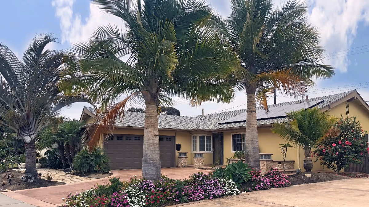 Exterior view of a single-story residential building with a beige facade, a two-car garage, and a tiled roof. The front yard features two tall palm trees, colorful flower beds, and various shrubs under a partly cloudy sky.
