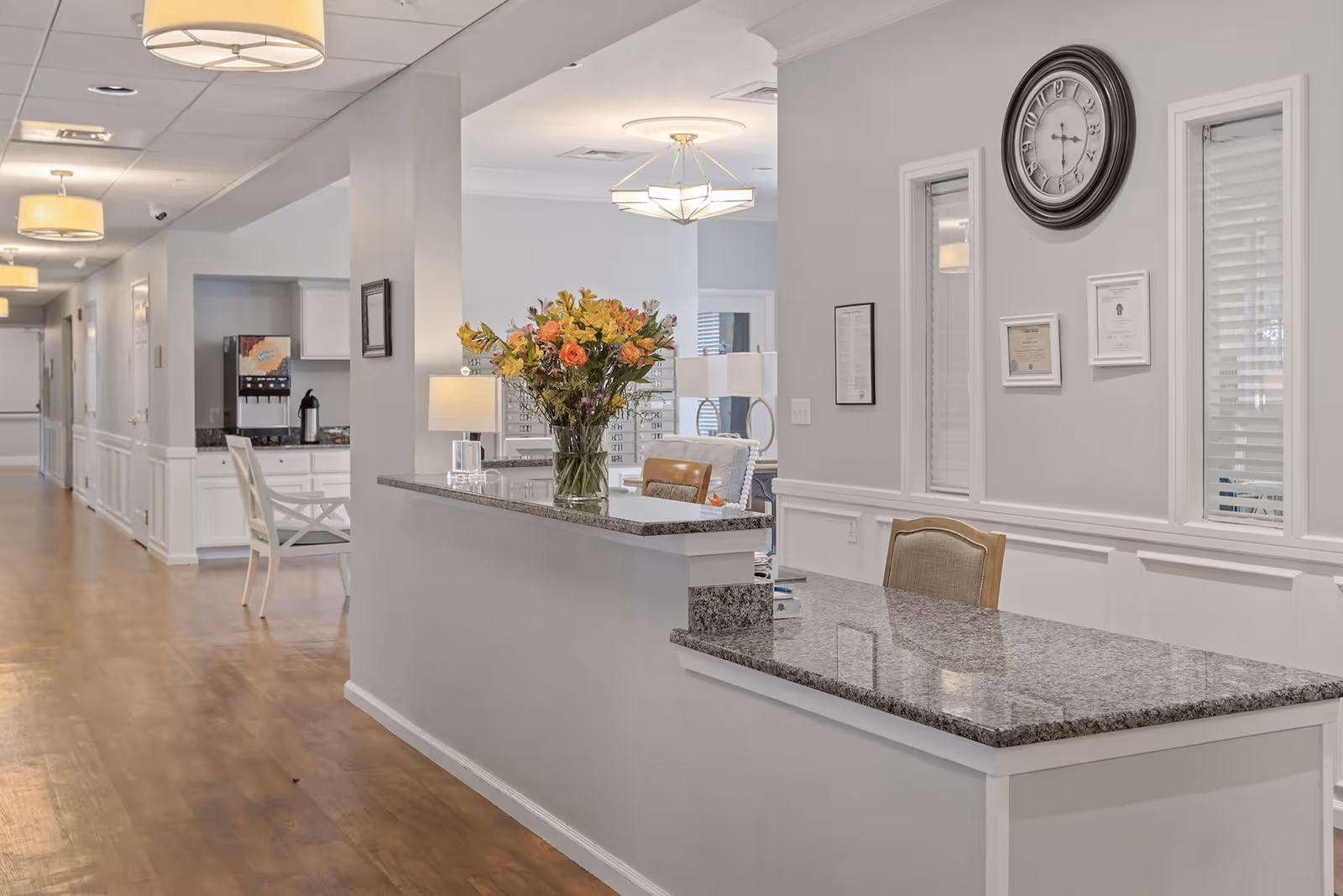 Interior view of a senior living facility reception area with a granite countertop desk, a vase of colorful flowers, a lamp, and a clock on the wall. The hallway extends to the left with wooden flooring and white walls, featuring multiple doors and ceiling lights.