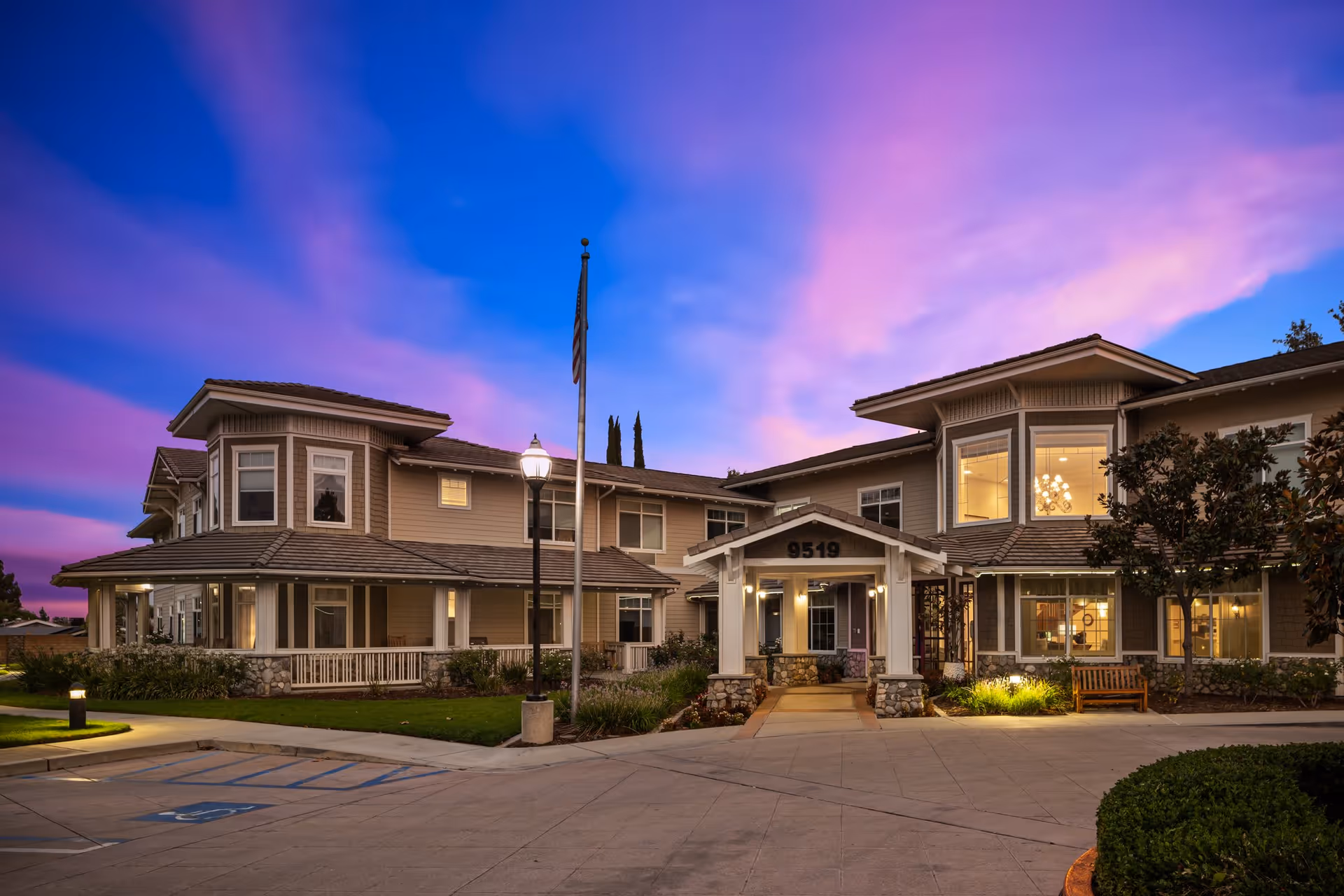 Exterior view of a two-story senior living facility building at dusk with a purple and blue sky. The building has beige siding, multiple windows, a covered entrance with the number 9519 above it, a flagpole with an American flag, and surrounding landscaping including bushes and trees. There is a parking area in front with a handicapped parking space visible.