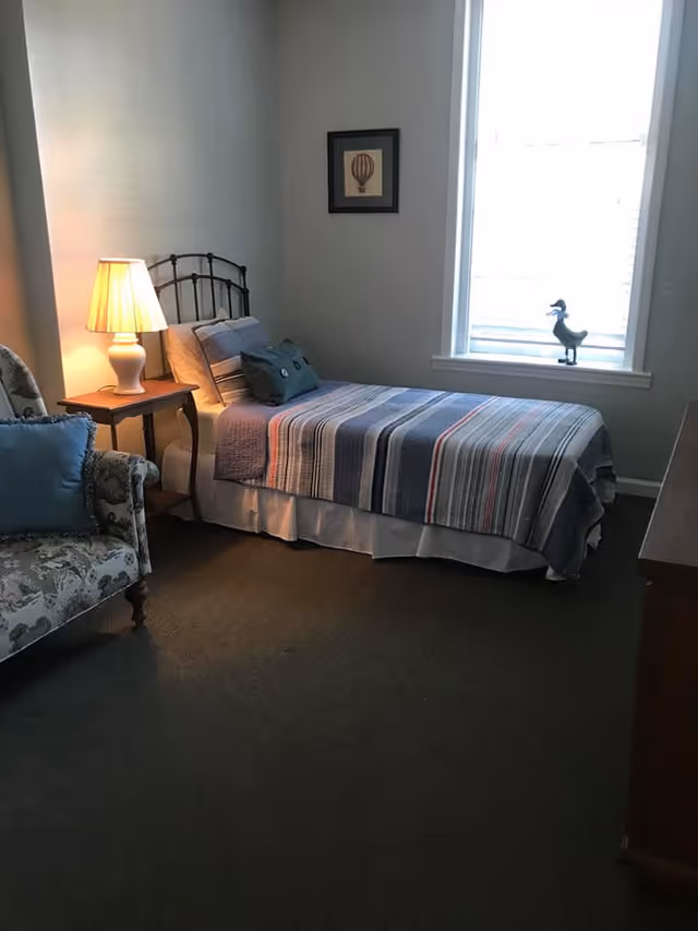 A small bedroom with a single bed covered in a striped quilt, a bedside table with a lamp, a patterned armchair with a blue cushion, a framed picture on the wall, and a window with a decorative duck figurine on the windowsill.