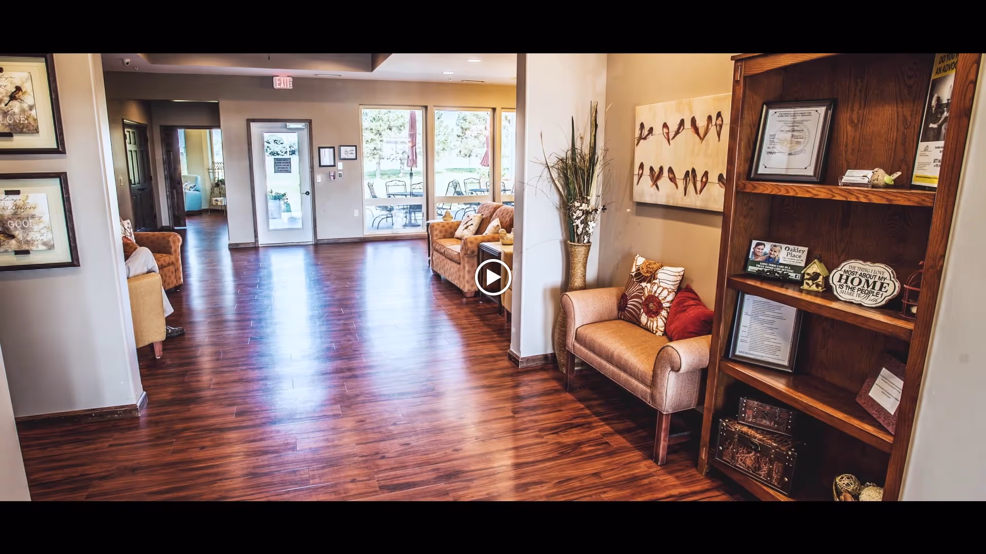 Interior view of a senior living facility hallway with polished wooden floors, beige walls, and comfortable seating areas with armchairs and cushions. A wooden bookshelf on the right holds framed certificates, decorative items, and brochures. Large windows and a glass door at the end of the hallway provide a view of an outdoor patio with tables and chairs.