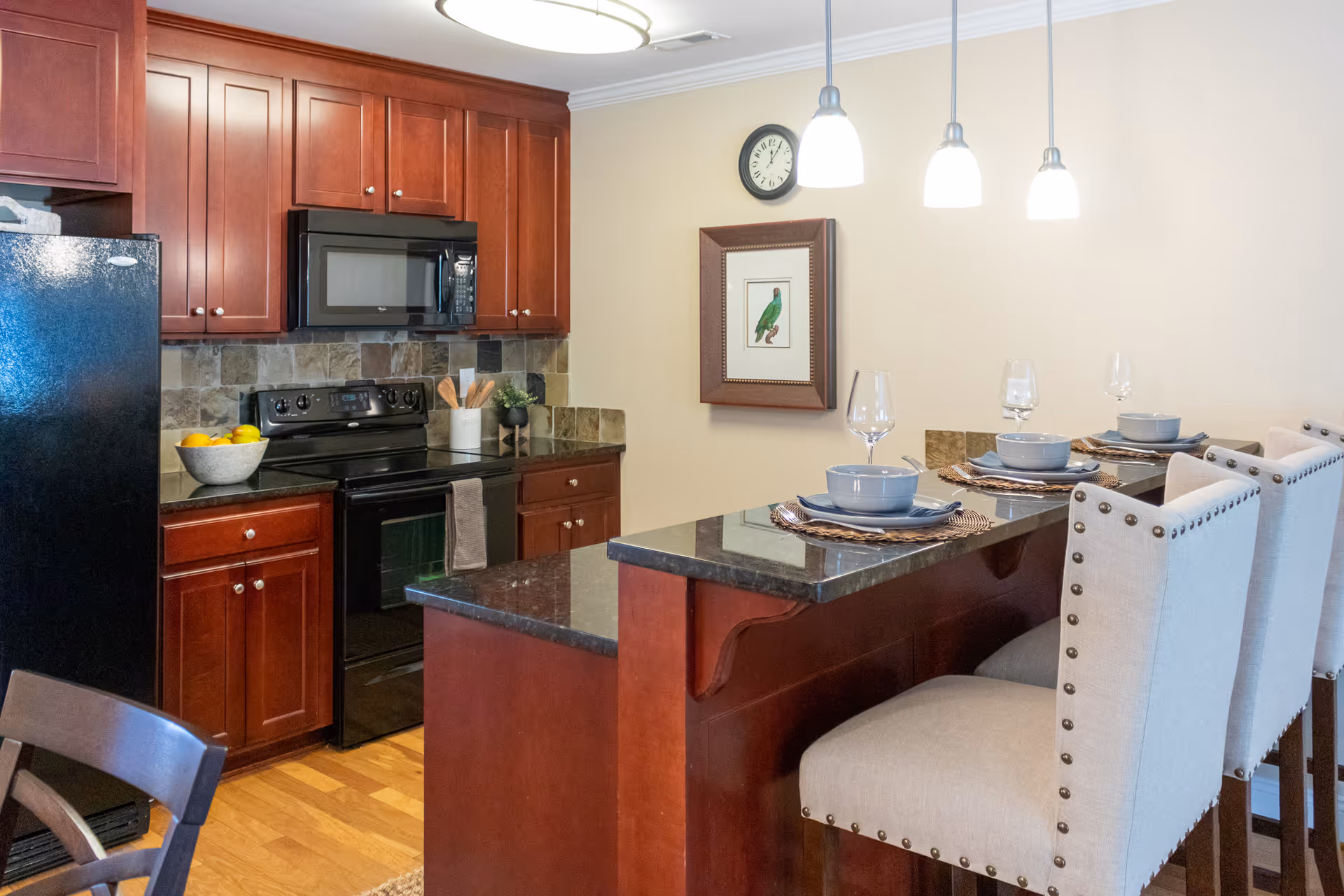 A modern kitchen with dark wood cabinets, black appliances including a refrigerator, stove, and microwave. There is a granite countertop with a breakfast bar featuring three beige upholstered bar stools with nailhead trim. The countertop is set with placemats, bowls, plates, and wine glasses. On the wall, there is a clock and a framed picture of a green bird. Three pendant lights hang above the breakfast bar.