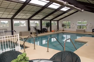 Indoor swimming pool area with clear water, surrounded by beige tiled flooring and metal handrails. The pool room has large windows letting in natural light and a high ceiling with skylights. There are some chairs and plants around the pool area.