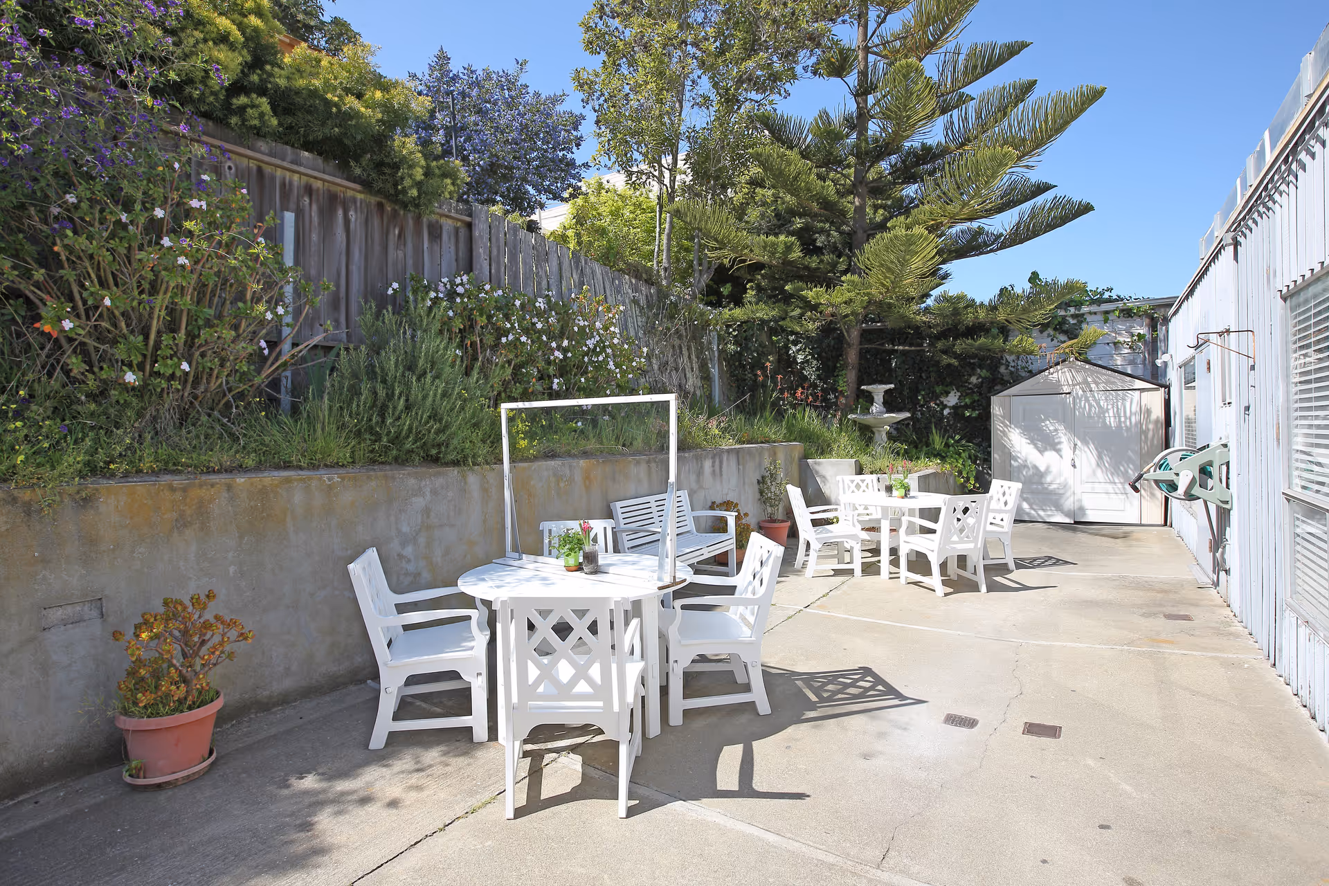 Outdoor patio area with white tables and chairs arranged on a concrete surface. There are plants and flowers along a raised garden bed with a wooden fence behind it. A small white shed and a birdbath are visible in the background under a clear blue sky.