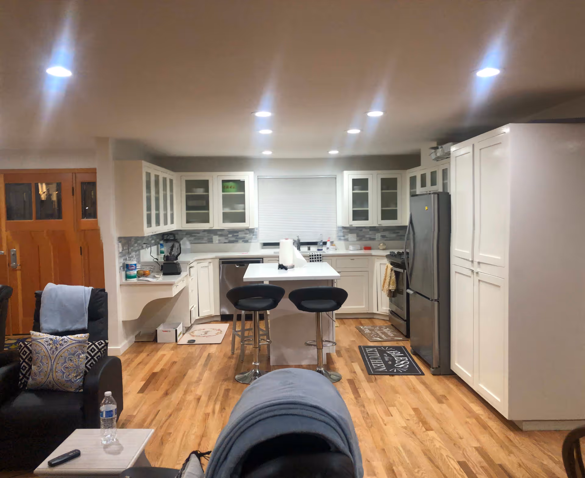 A modern kitchen with white cabinetry, a central island with two black bar stools, stainless steel refrigerator and dishwasher, wooden flooring, and recessed ceiling lights. To the left, part of a living area with a black armchair, pillows, and a small table with a water bottle is visible.