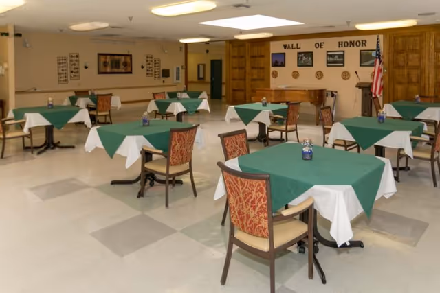 A dining room with multiple square tables covered with green and white tablecloths. Each table has two chairs with patterned upholstery. The room has a tiled floor, beige walls, and ceiling lights. At the far end, there is a wooden wall with the words 'Wall of Honor,' several framed pictures, plaques, and an American flag beside a piano.