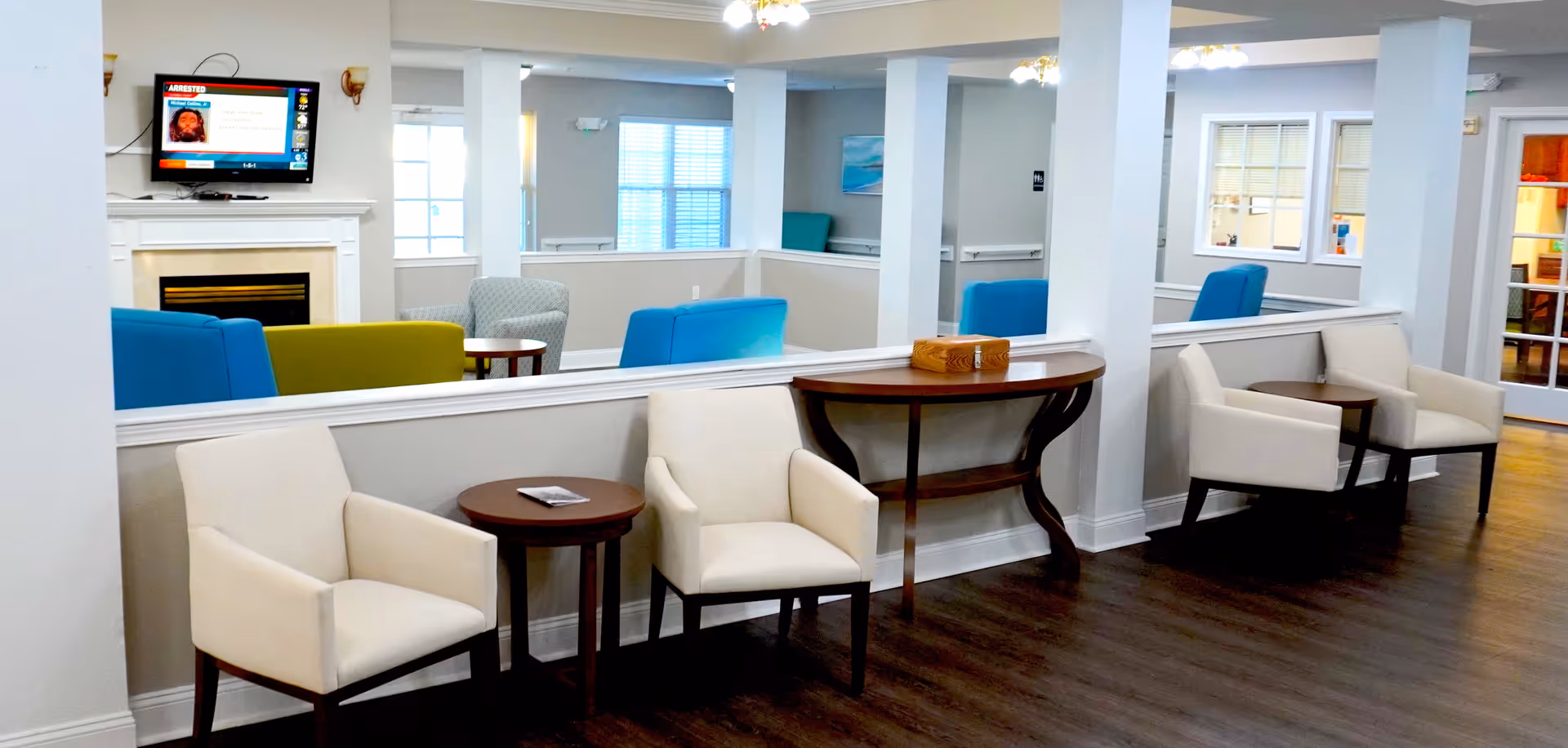 A bright and clean common area in a senior living facility with cream-colored armchairs, small wooden tables, a fireplace with a mounted TV above it, and colorful seating in the background. The room has large windows with blinds and light gray walls.