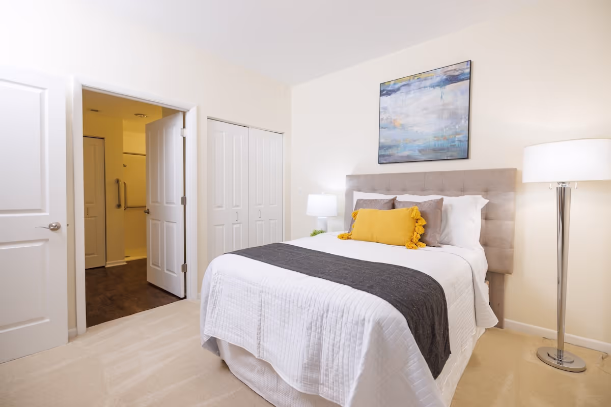 A neatly made bed with white bedding, a dark gray runner, and decorative pillows including a yellow one with tassels in a bright bedroom. There is a floor lamp to the right of the bed and a framed abstract painting on the wall above the headboard. To the left, there is an open door leading to a bathroom and a closed closet with double doors.