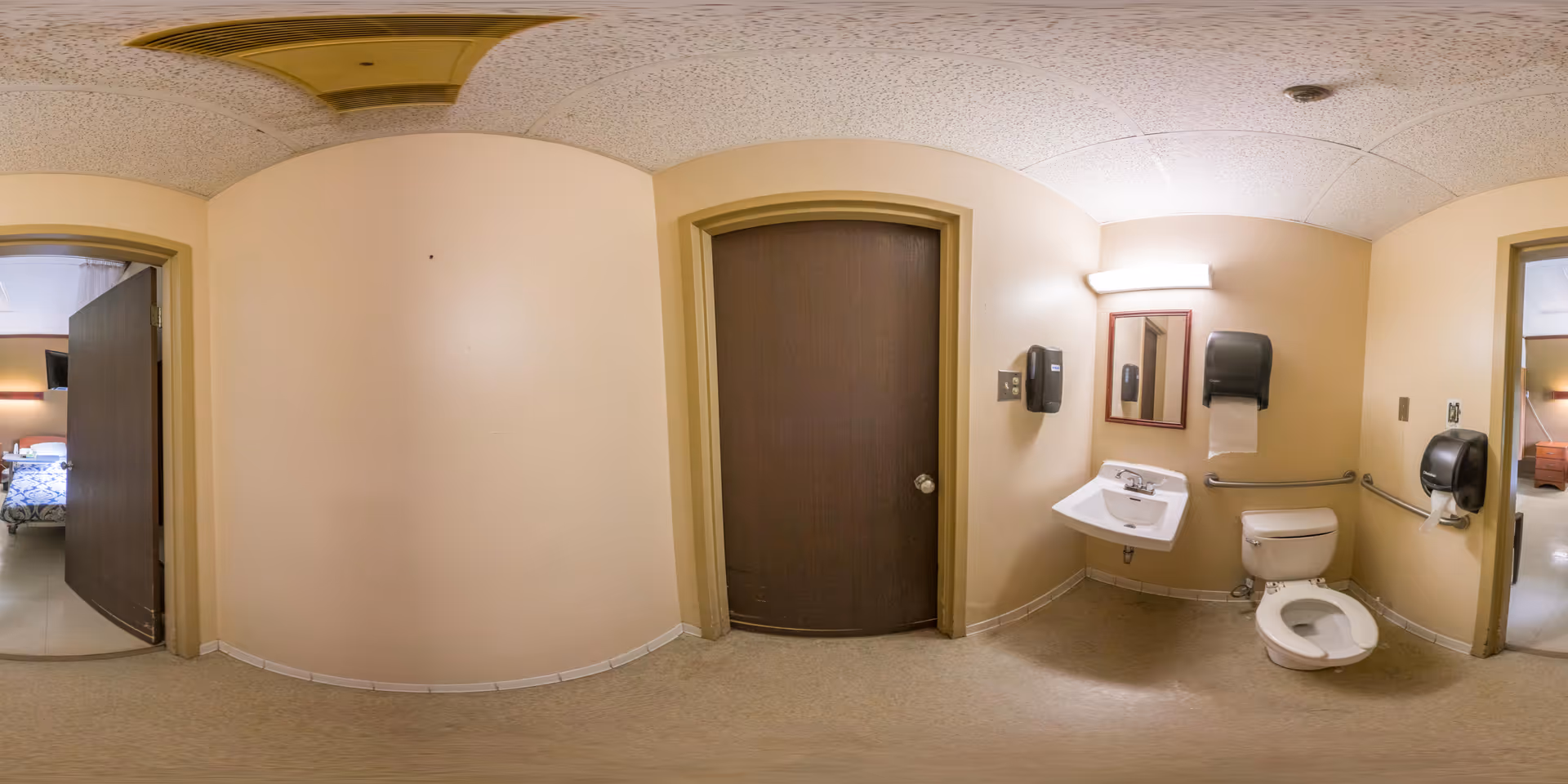 Interior view of a nursing facility bathroom with beige walls and tiled floor. The bathroom includes a white toilet with a safety grab bar, a white sink with a mirror and a light above it, and two black dispensers mounted on the walls. There are two doorways visible, one leading to a bedroom with a bed and TV, and the other doorway is closed with a dark brown door.