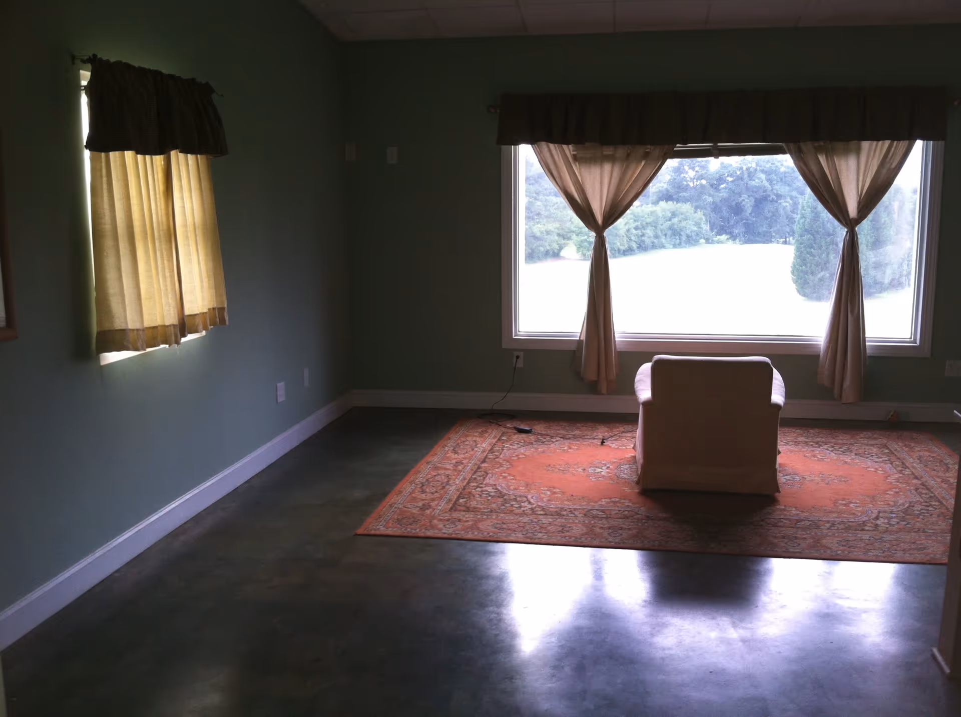 Sparsely furnished living room with a single armchair on an orange rug in front of a large window with curtains.