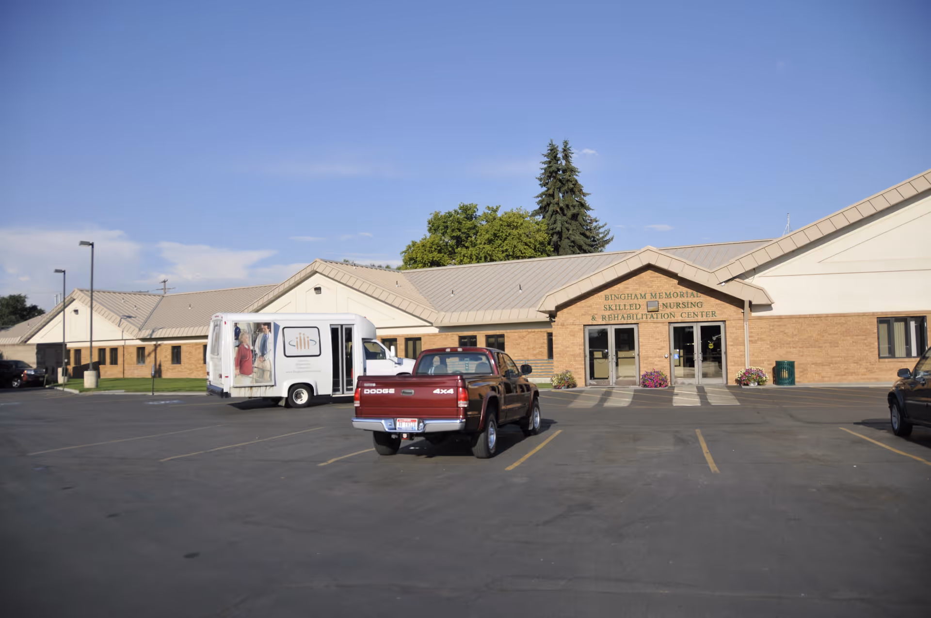 Exterior view of Bingham Memorial Skilled Nursing and Rehabilitation Center building with a parking lot in front. Several vehicles, including a red Dodge truck and a white van with images of people, are parked. The building has a beige brick facade and a light-colored roof under a clear blue sky.
