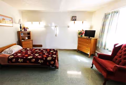 A furnished senior living bedroom with a bed, dresser, TV, and red armchair near a window.