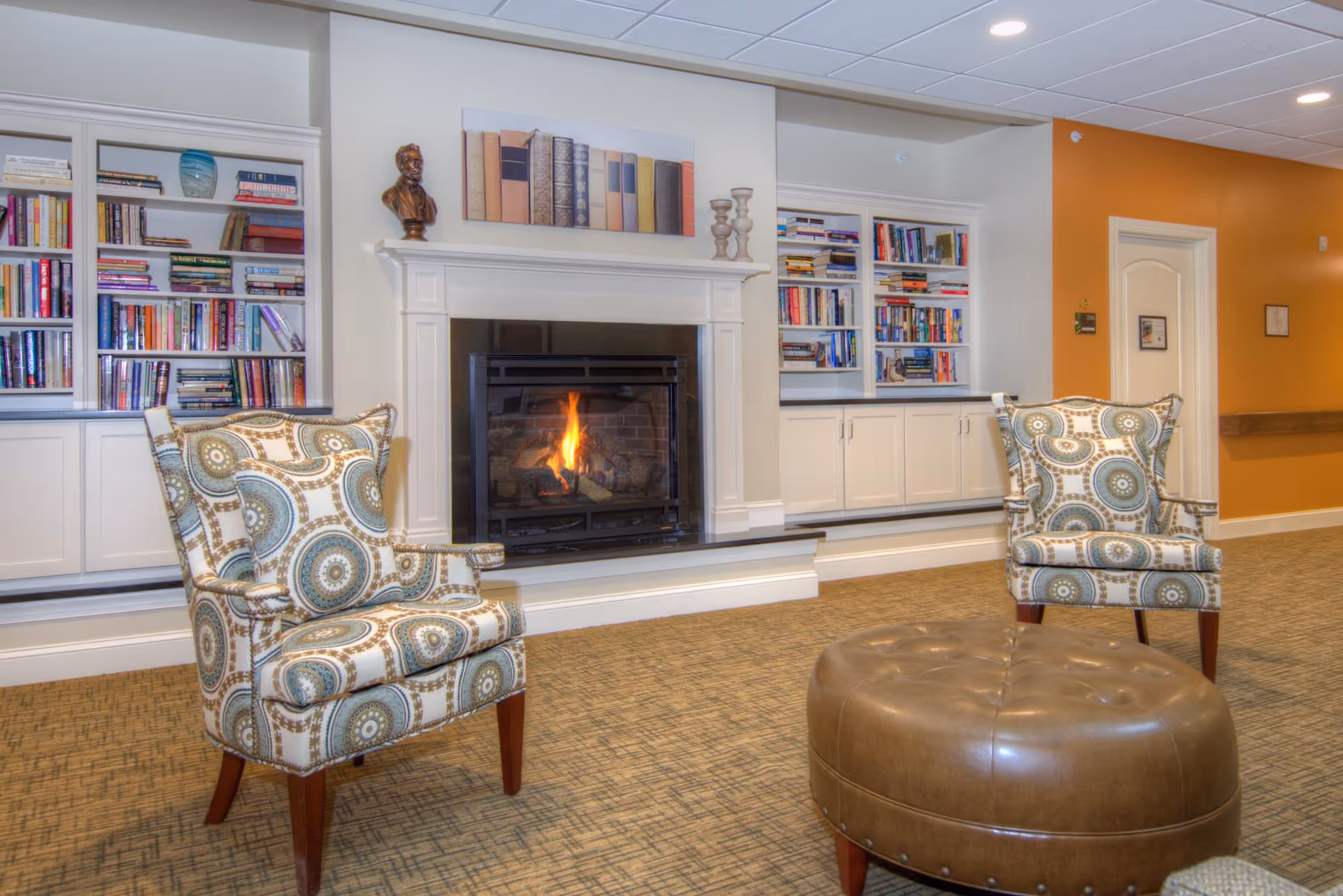 A cozy senior living common area with two patterned armchairs facing a round leather ottoman. Behind the chairs is a lit fireplace framed by built-in bookshelves filled with books and decorative items. The walls are painted white and orange, and the carpet has a textured pattern.
