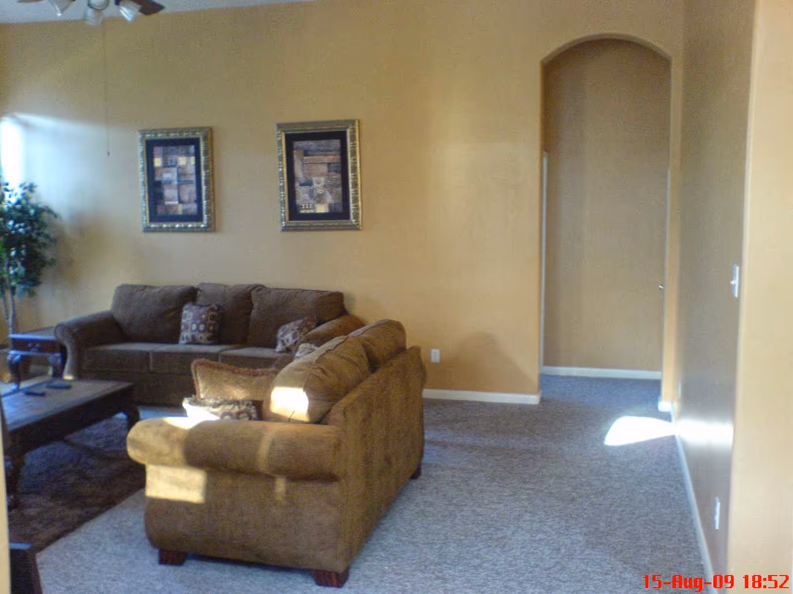 A living room with two brown sofas arranged around a wooden coffee table. The walls are painted a warm beige color and decorated with two framed abstract art pieces. There is a potted plant in the corner near a window letting in natural light. The floor is carpeted in a light gray tone, and there is an arched doorway leading to another room.