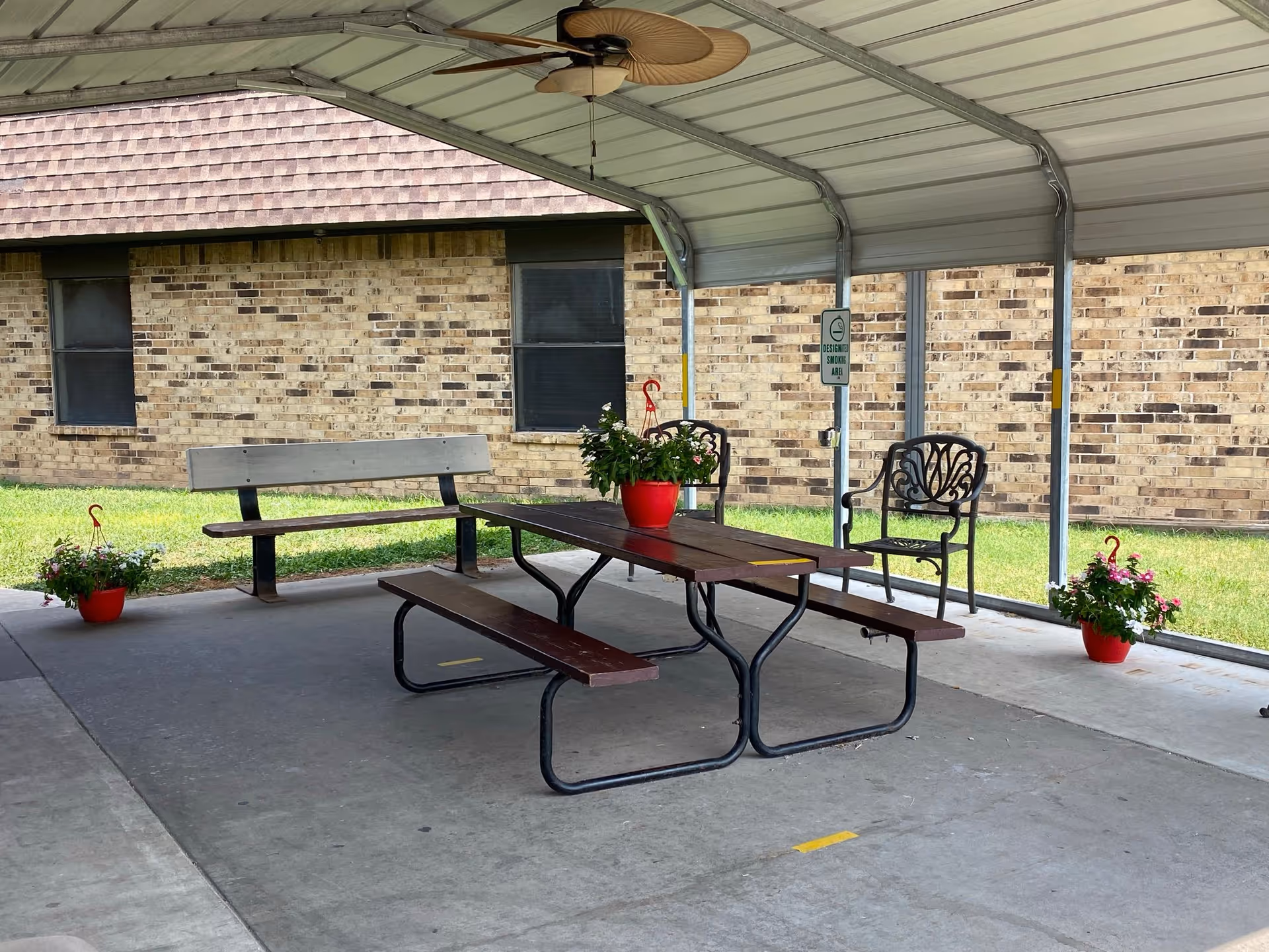 Covered outdoor seating area with a picnic table, benches, and chairs. There are potted plants with flowers on and around the table. The area is next to a brick building with two windows and a grassy lawn.