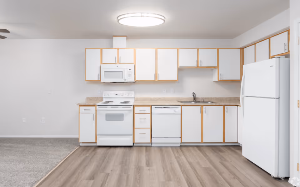 A clean and modern kitchen with white cabinets and light wood trim, featuring a white stove with an overhead microwave, a dishwasher, a double sink, and a white refrigerator. The kitchen has light wood flooring and is adjacent to a carpeted area.