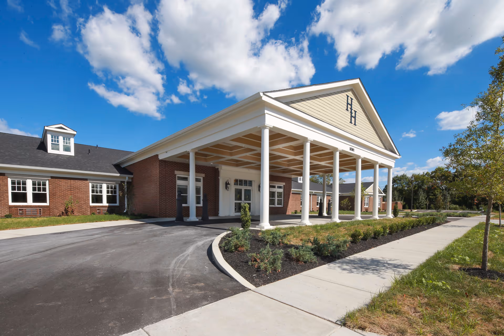 Exterior view of Hallmark House senior living facility showing a large covered entrance with white columns, brick walls, and a clear blue sky with some clouds.