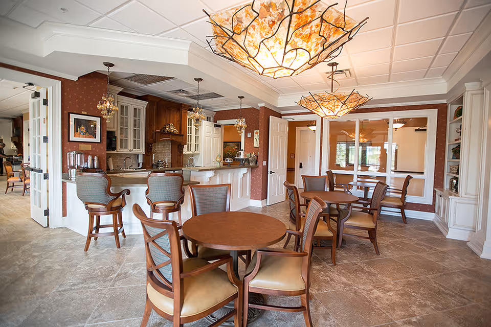 A cozy dining area in a senior living facility featuring round wooden tables with cushioned chairs. The room has a warm color palette with brown walls and beige tiled flooring. There is a kitchen area with a counter and bar stools in the background, and decorative pendant lights hang from the ceiling. Large windows and glass doors allow natural light to brighten the space.
