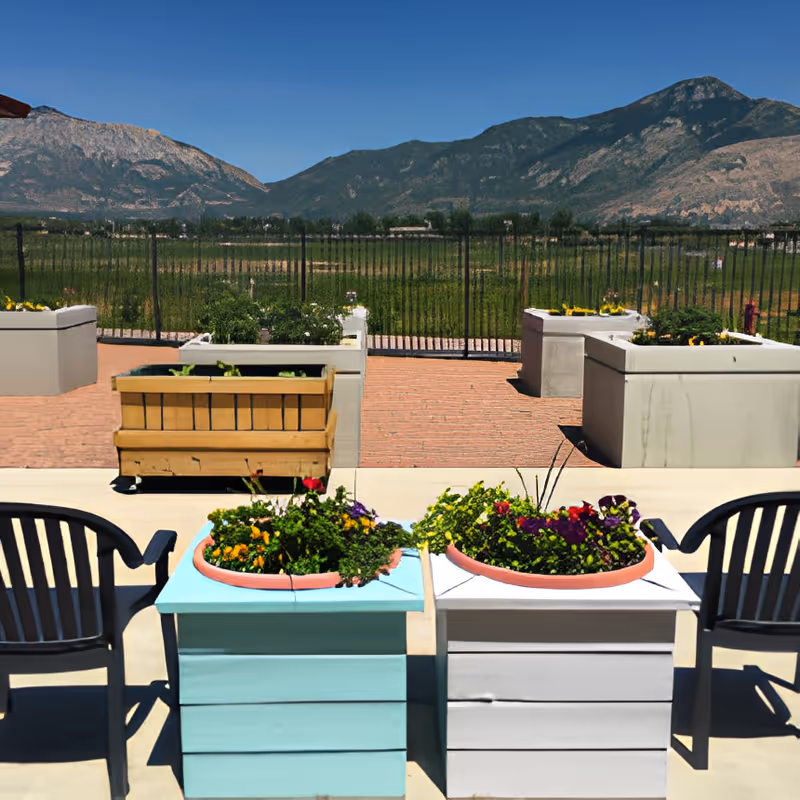 Outdoor patio area with flower planters and two black chairs facing a fenced green field with mountains in the background under a clear blue sky.