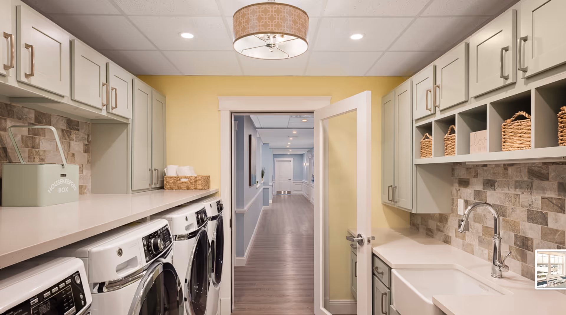 Bright laundry room with multiple front-loading washers and dryers, sink, cabinetry, and a hallway visible through an open door.