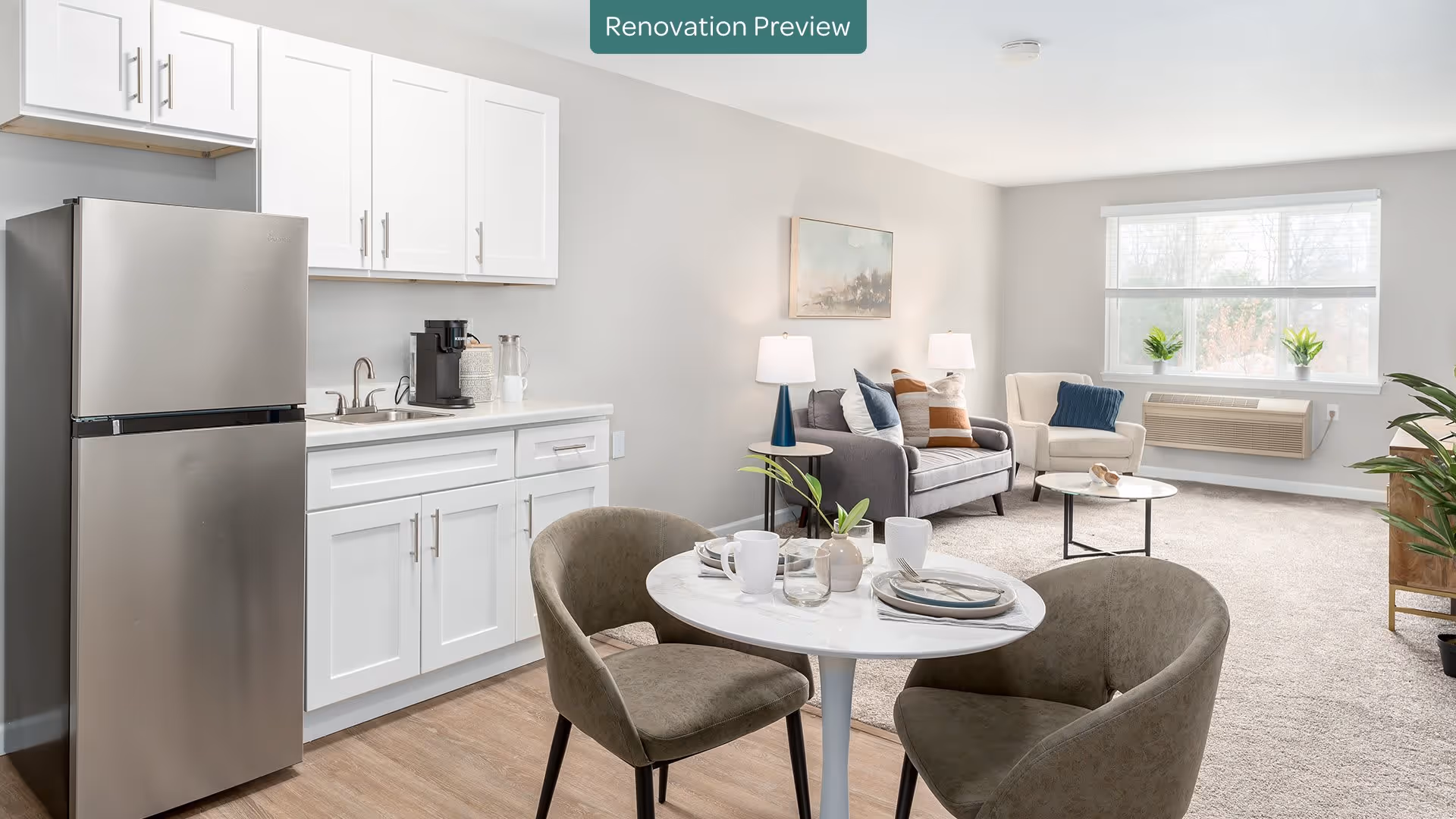 A bright and modern living space featuring a small kitchen area with white cabinets, a stainless steel refrigerator, and a coffee maker on the counter. In front of the kitchen is a round white dining table set with plates, cups, and utensils, surrounded by two upholstered chairs. The living area includes a gray sofa with colorful pillows, a white armchair, two table lamps, a small round coffee table, and a large window letting in natural light. The room has light-colored walls and carpeted flooring.