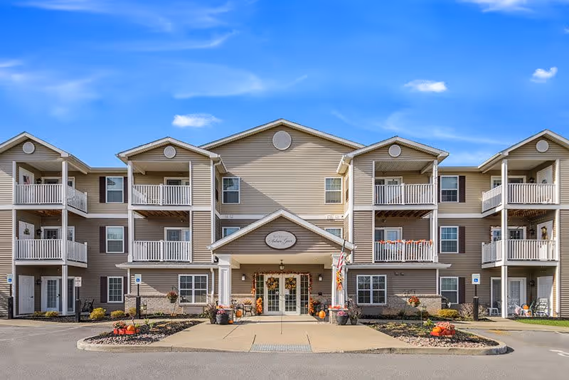 Front exterior view of a three-story beige apartment building with multiple balconies and a central entrance decorated with fall-themed wreaths and pumpkins under a clear blue sky.