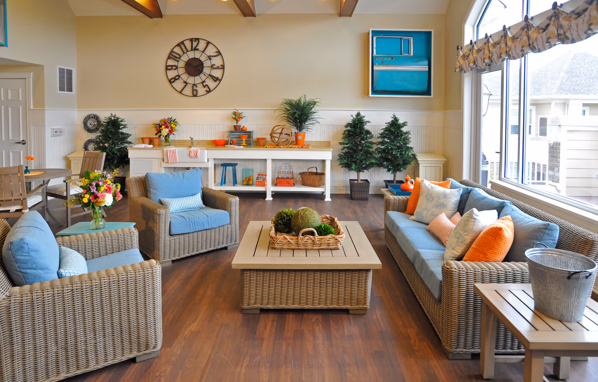 Bright communal living room with wicker sofas and armchairs, a central coffee table, decorative pillows and plants, and large windows.