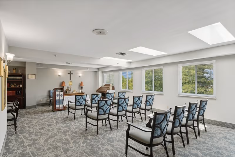A small chapel or prayer room with rows of chairs facing a small altar with a cross on the wall behind it. The room has large windows letting in natural light and a patterned carpet floor.