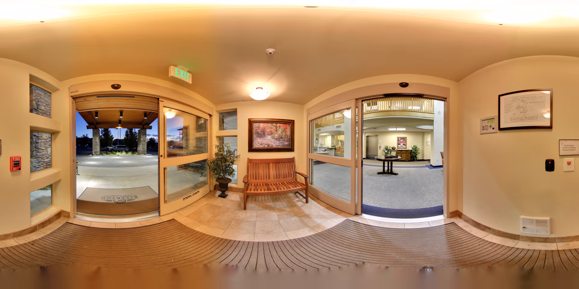 Entrance area of a senior living facility with automatic sliding glass doors leading outside and inside. There is a wooden bench with a potted plant next to it, a framed picture on the wall, and an exit sign above the door. The interior visible through the right door shows a reception area with a table and plants.