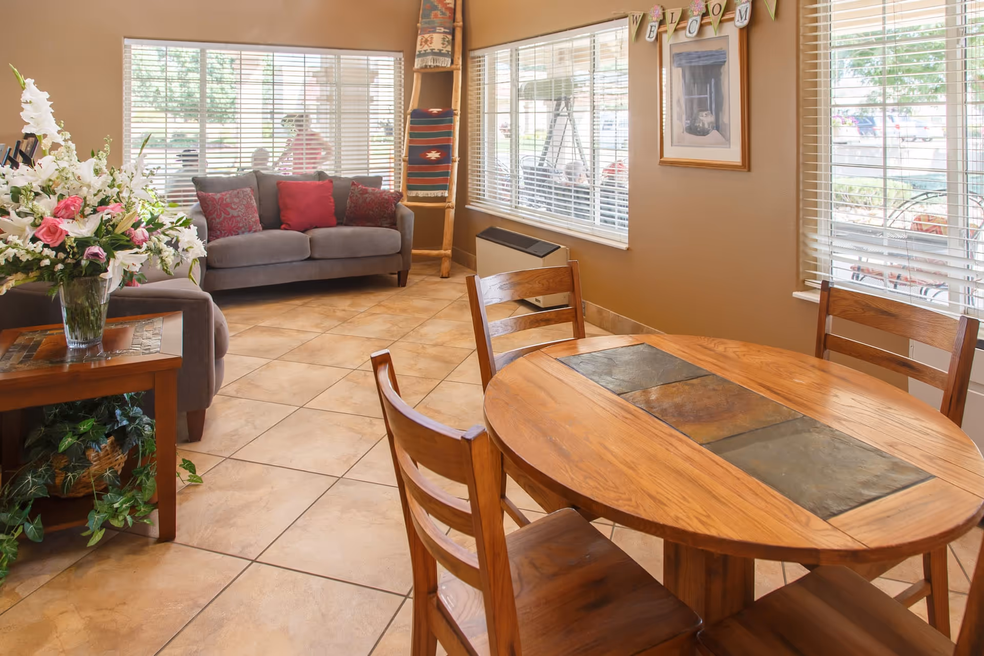 Sunny common area with a wooden round dining table and chairs, tiled floor, and a sofa by large windows.
