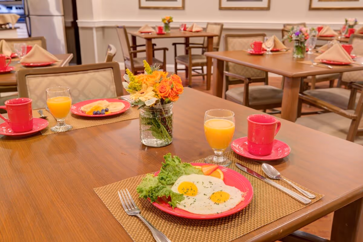 A dining room with wooden tables set for breakfast. One table in the foreground has a plate with two sunny-side-up eggs, lettuce, and fruit, along with a glass of orange juice and a red coffee cup on a saucer. Other tables in the background are similarly set with red plates, cups, and folded napkins. A small vase with orange and yellow flowers is on the table in the foreground.
