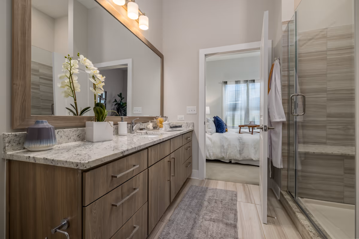 Modern bathroom with a large mirror above a granite countertop vanity with wooden cabinets. There is a white orchid plant and other small decorative items on the counter. To the right, there is a glass-enclosed shower with beige tiles. The bathroom door is open, showing a bedroom with a bed, blue pillows, a side table, and a window with blinds and curtains.
