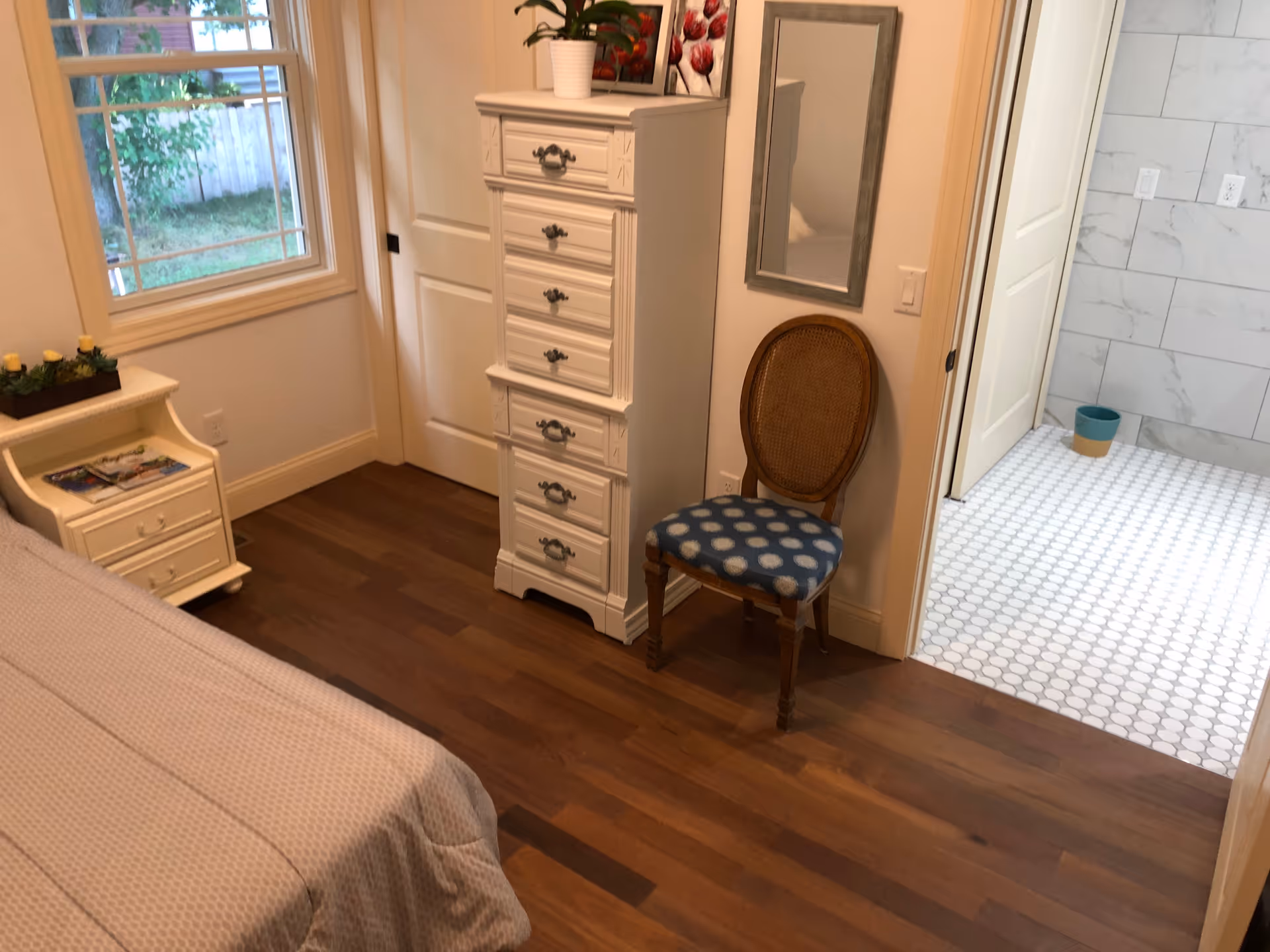 A cozy bedroom corner with wooden flooring, a white tall chest of drawers, a wooden chair with a blue polka dot cushion, and a small white nightstand with magazines and a decorative plant. A window shows greenery outside. An open door leads to a bathroom with white hexagonal floor tiles and marble-patterned wall tiles.