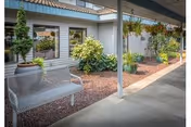 Covered exterior walkway with a metal bench, potted and hanging plants, and windows along the building.