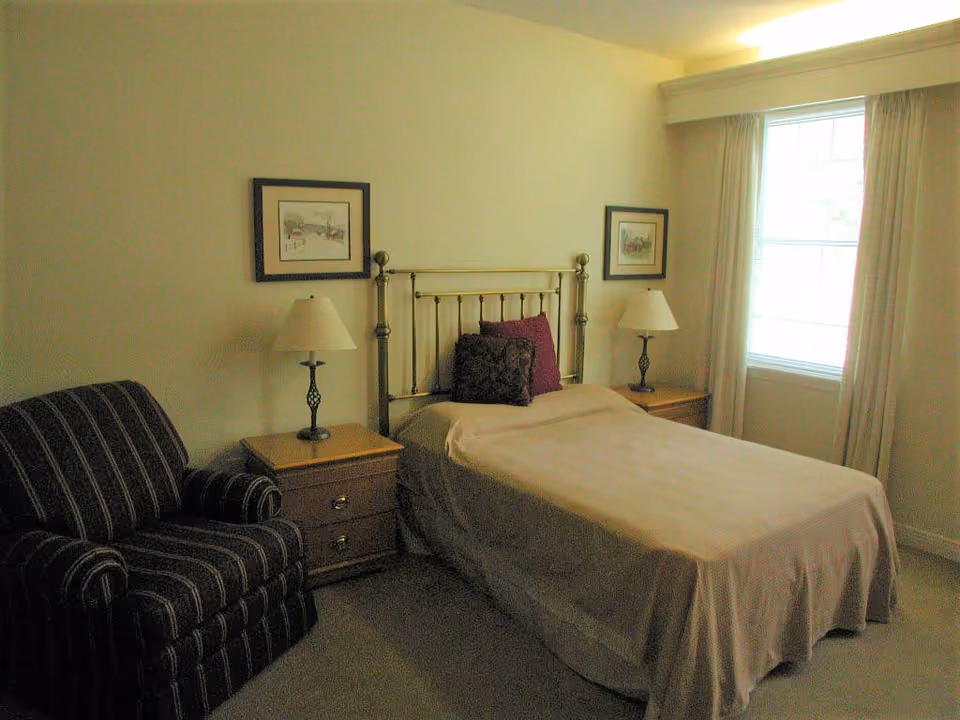 A bedroom with a single bed covered in a beige bedspread, two pillows, and a metal headboard. There are two wooden nightstands on either side of the bed, each with a lamp. A striped armchair is positioned to the left of the bed. The room has light-colored walls, a window with curtains on the right side, and two framed pictures hanging above the nightstands.