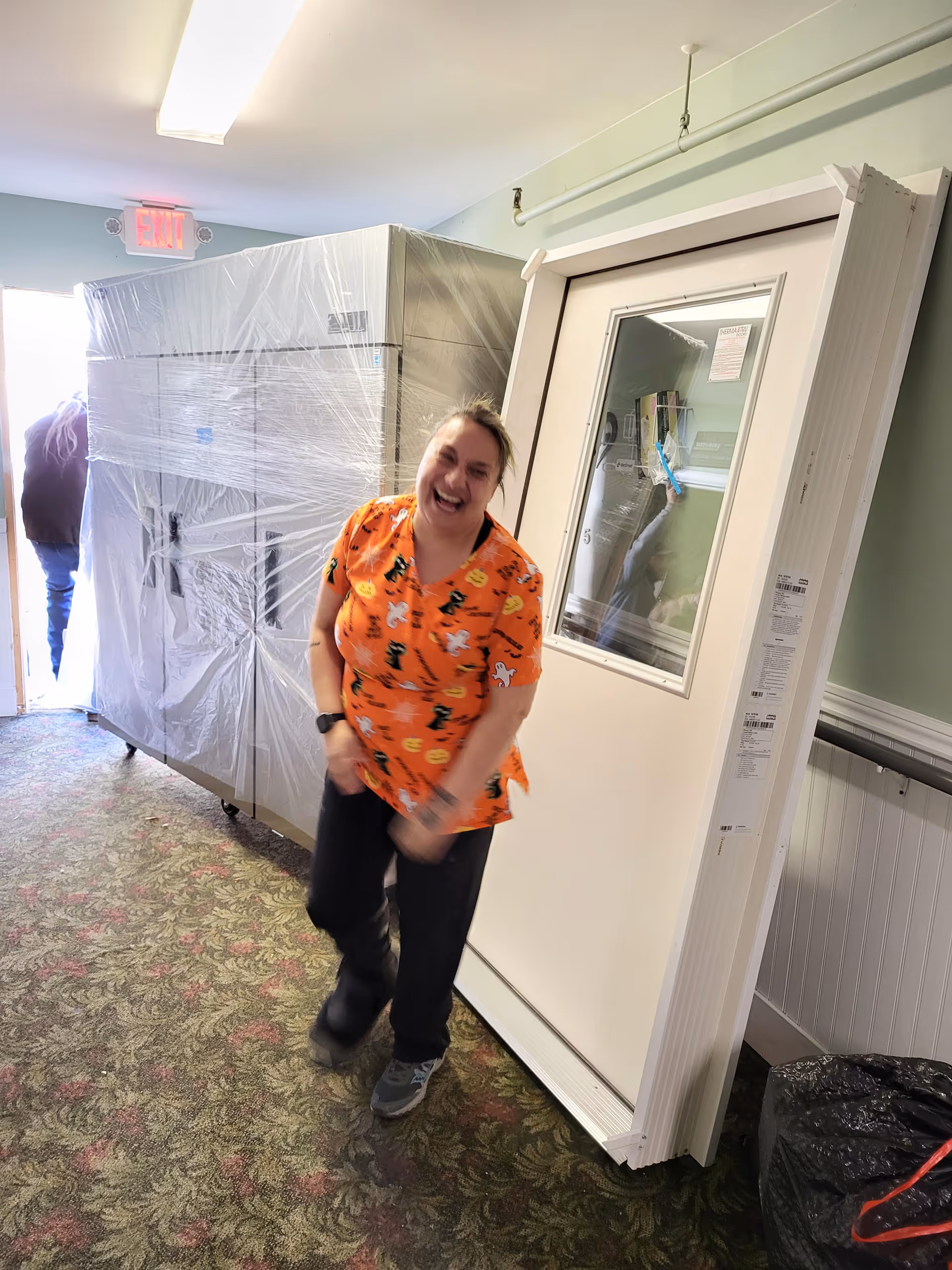 A woman wearing an orange Halloween-themed shirt with black pants is laughing and walking down a carpeted hallway. Behind her, there is a large appliance wrapped in plastic and a white door with a window leaning against the wall. Another person is partially visible near an exit door at the end of the hallway.