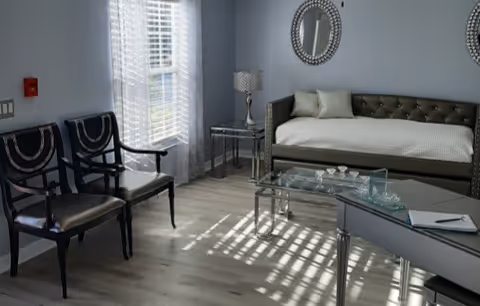 A bright room with light gray walls and wood flooring featuring two black chairs with decorative backs, a glass coffee table, a gray daybed with cushions, a side table with a lamp, and two round mirrors on the wall. Sunlight streams through a window with white blinds and sheer curtains.
