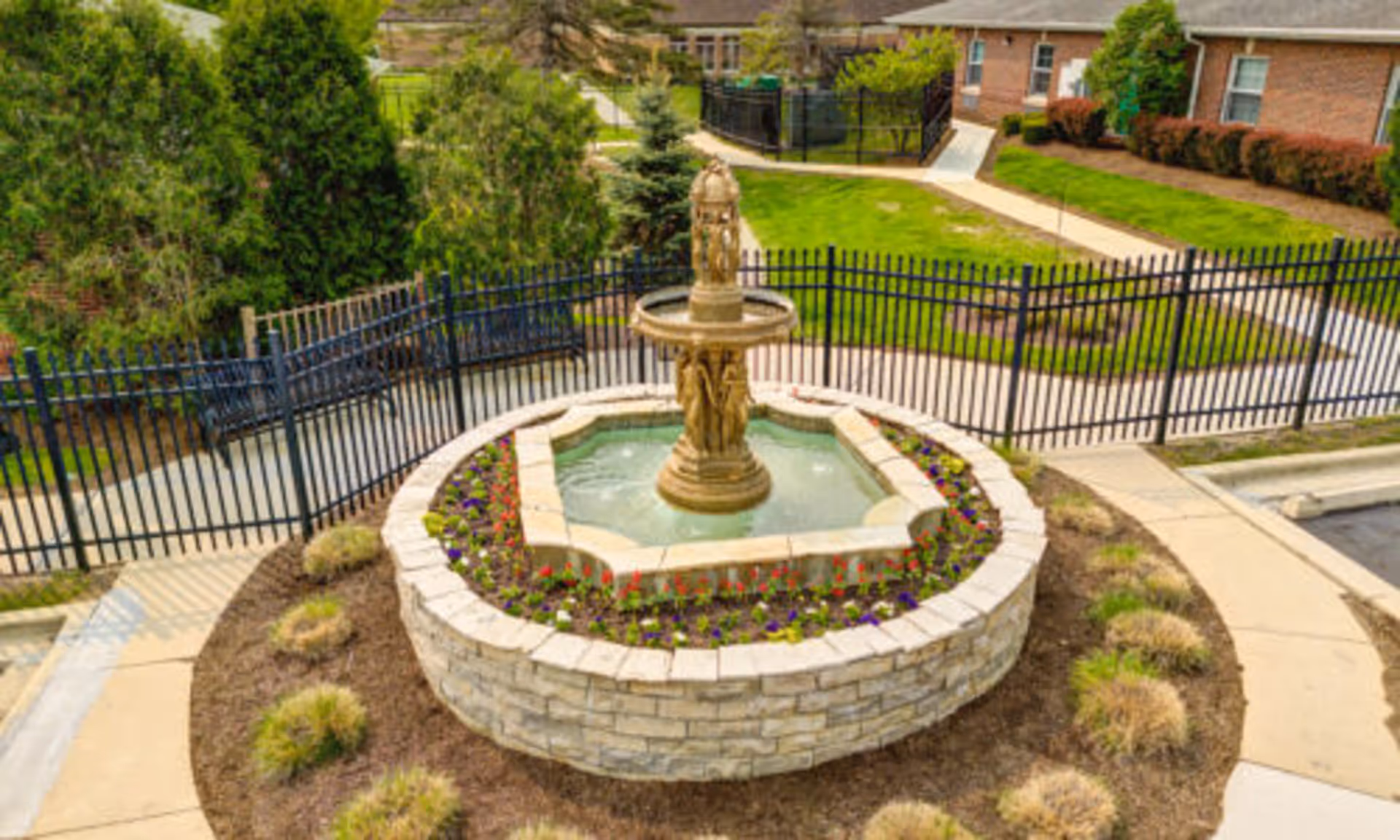 A stone tiered fountain in the center of a circular flowerbed in a fenced landscaped courtyard with walkways and brick buildings in the background.