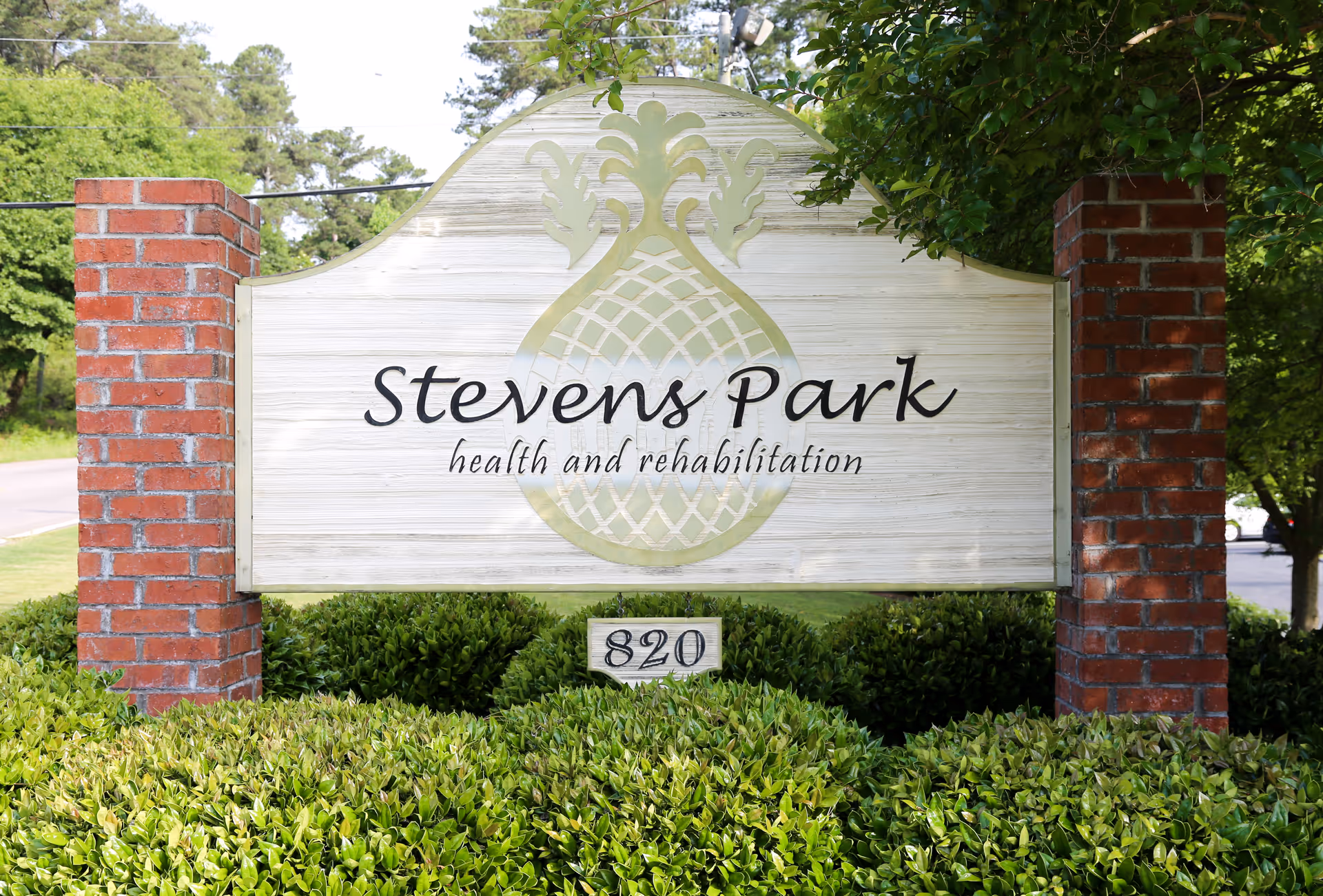 A large outdoor sign for Stevens Park health and rehabilitation, mounted between two red brick pillars with green bushes in front and trees in the background.